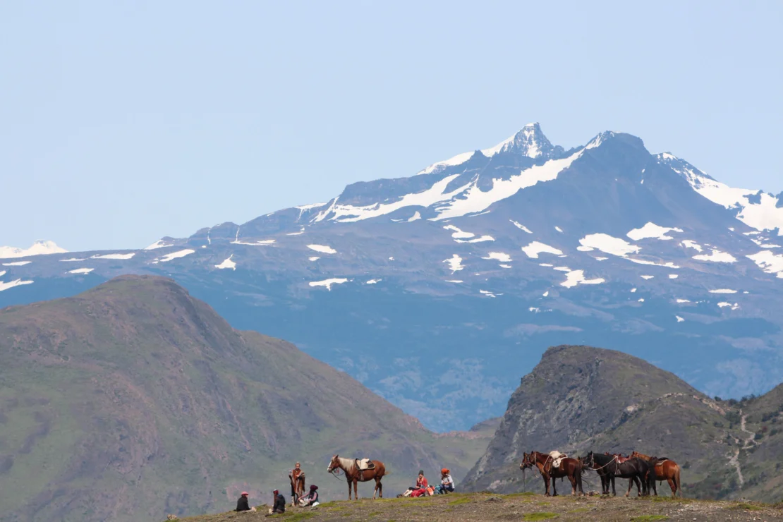 People and horses rest on a grassy hillside with snow-capped mountains in the background under a clear sky.