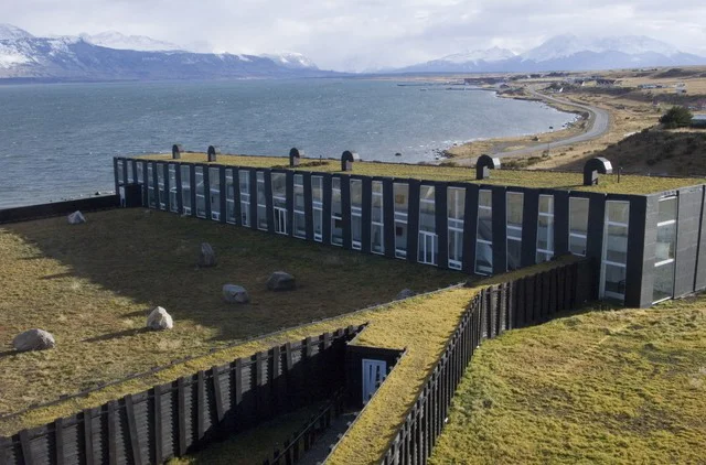 A long, rectangular building with a grass-covered roof is situated near a large body of water, with mountains in the background.