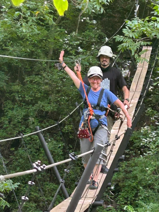 Two people wearing helmets and harnesses cross a narrow wooden suspension bridge in a forested area. The person in front waves while walking.