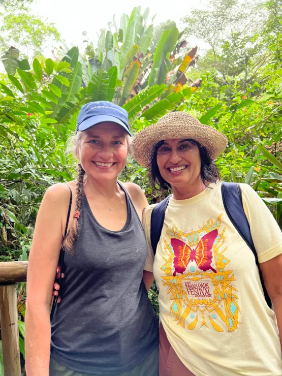 Two women smiling in front of lush green foliage. One wears a blue cap and tank top, the other a hat and butterfly-themed t-shirt.