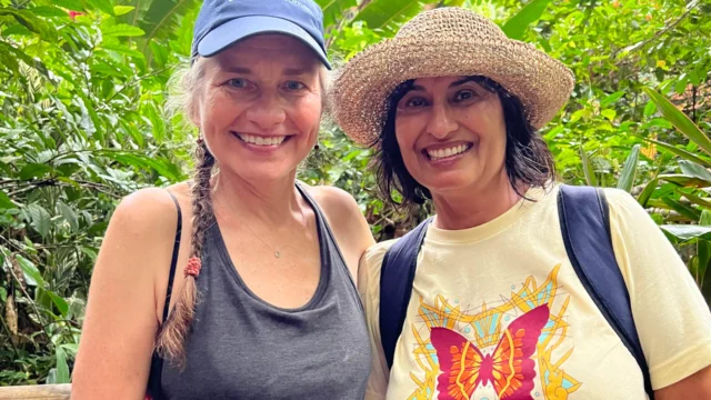 Two women smiling in front of lush green foliage. One wears a blue cap and tank top, the other a hat and butterfly-themed t-shirt.