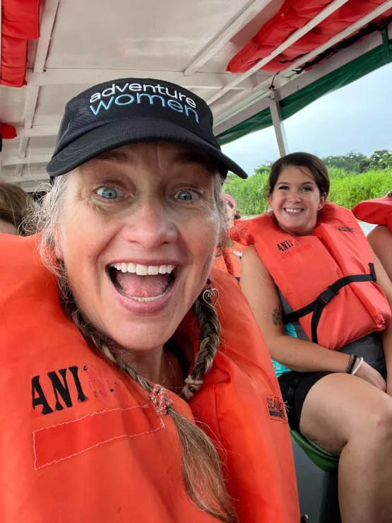 Two people wearing orange life jackets smile on a boat. One wears a cap with the text "adventure women.