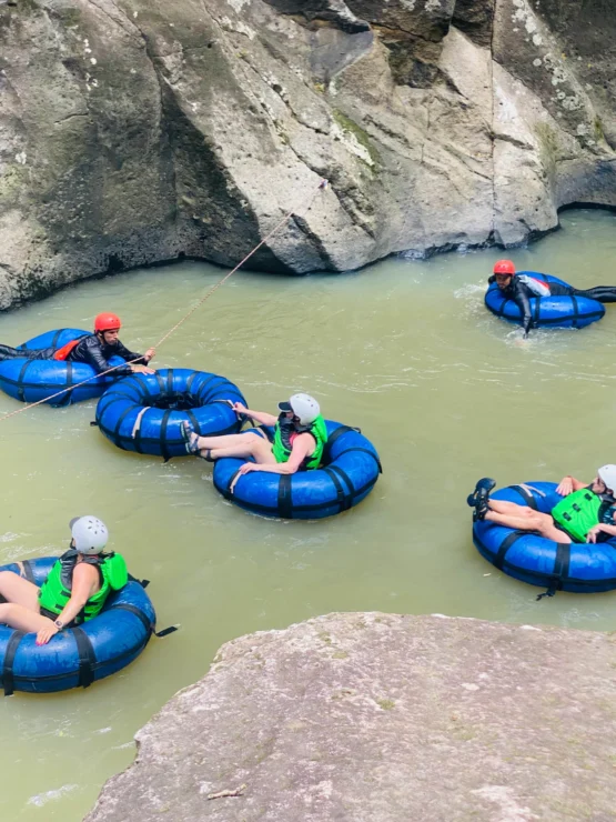 People in helmets and life vests are floating on blue inner tubes in a rocky river.