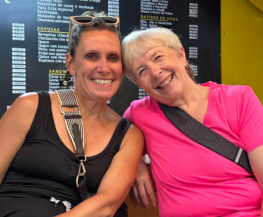 Two women smiling, sitting together in front of a menu board. One wears sunglasses on her head, and the other wears a pink shirt.