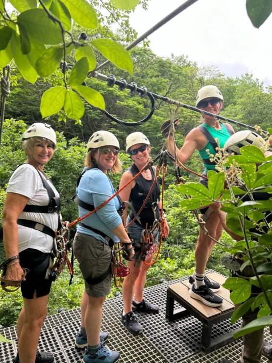 Four people in helmets and harnesses stand on a zipline platform in a lush forest setting.