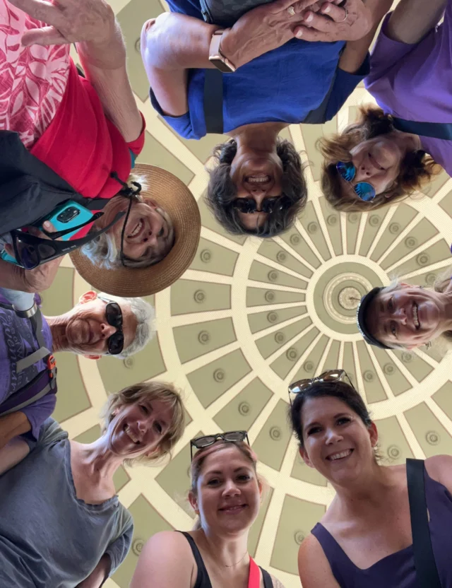 A group of people stand in a circle, looking down at the camera. Above them is a decorative domed ceiling.