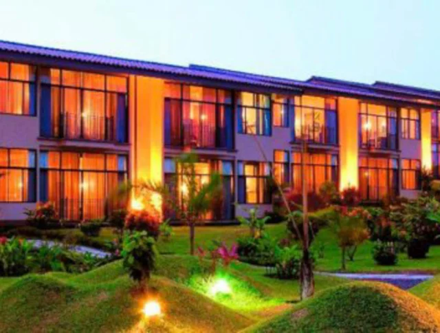 A modern, two-story building with large windows and balconies, illuminated by warm lights. It is surrounded by a well-kept garden with small hills and palm trees.