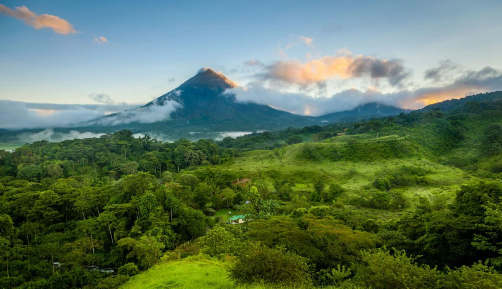 Mountain with clouds and forested landscape below, under a partly cloudy sky at sunrise or sunset.