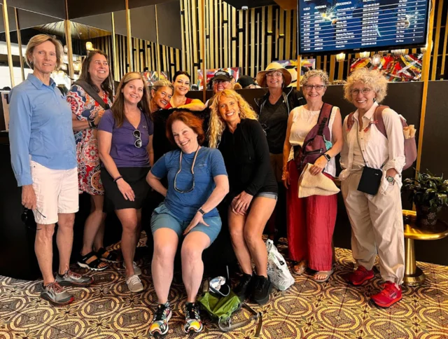 A group of ten people posing together indoors with a patterned carpet and a digital screen in the background.