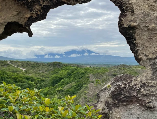 View of a distant mountain framed by a natural rock window, with lush green landscape and cloudy sky.