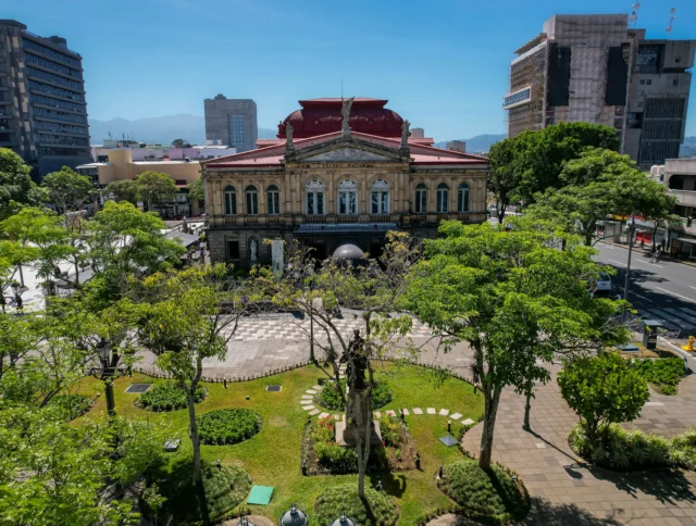 A historic building with a red roof and ornate facade is surrounded by trees and greenery in an urban setting.