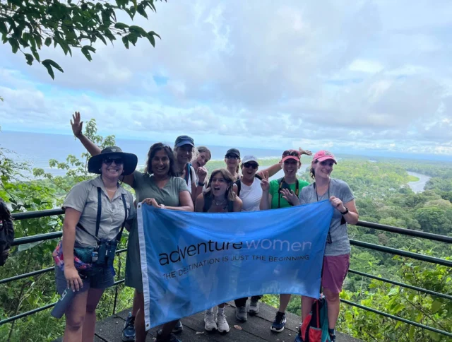 A group of people holding an "adventure women" banner pose for a photo on a scenic overlook, with lush greenery and the ocean in the background.