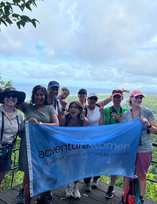 A group of people holding an "adventure women" banner pose for a photo on a scenic overlook, with lush greenery and the ocean in the background.