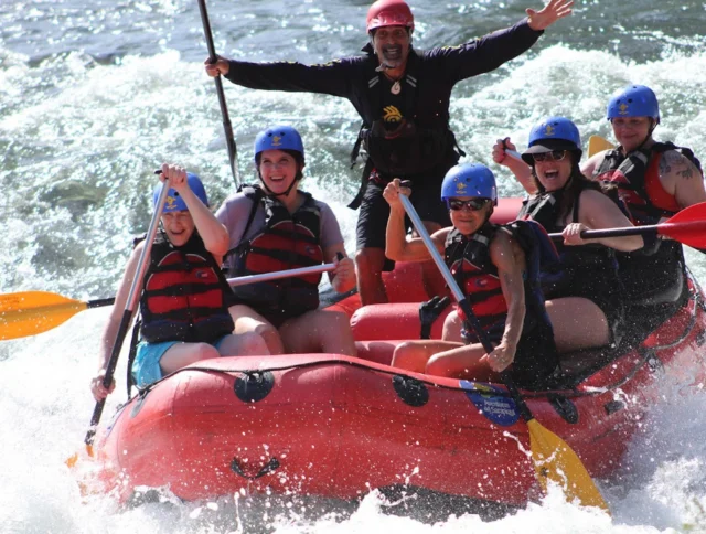 A group of six people wearing helmets and life vests joyfully rafting on a river, with splashes of water surrounding them.