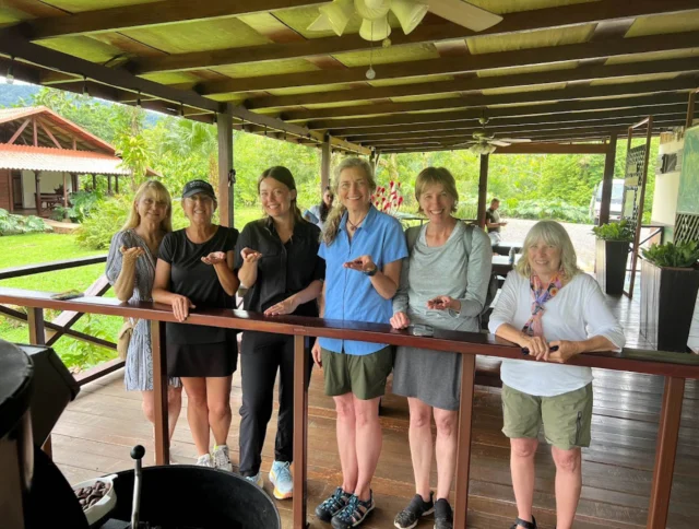 Six women stand on a wooden deck, holding objects in their hands, with greenery in the background.