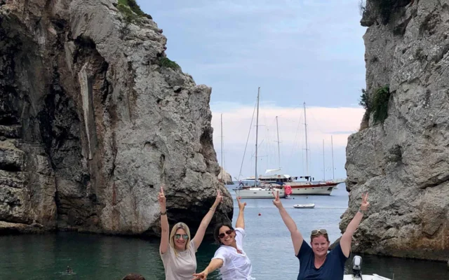 Four people pose energetically on a rocky beach with tall cliffs and anchored boats in the background.