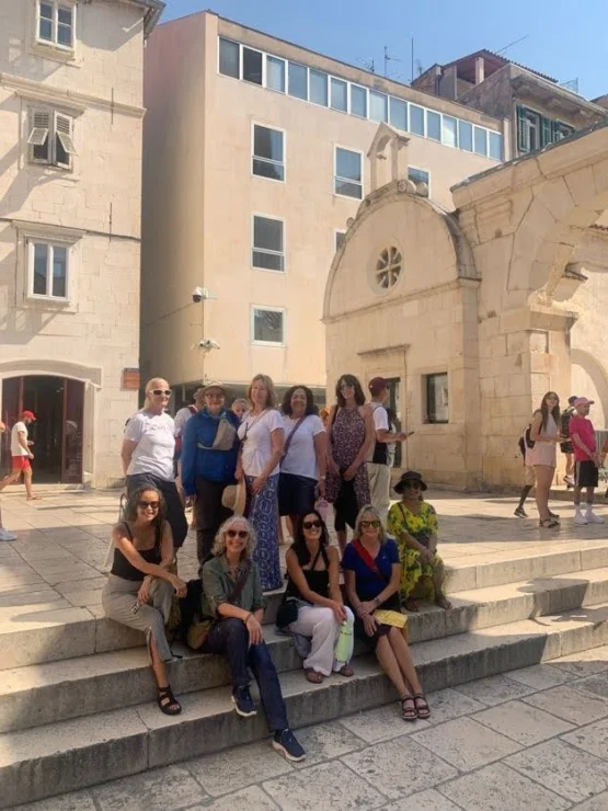 A group of people sitting on steps in front of a historic building with arches, under a clear sky.