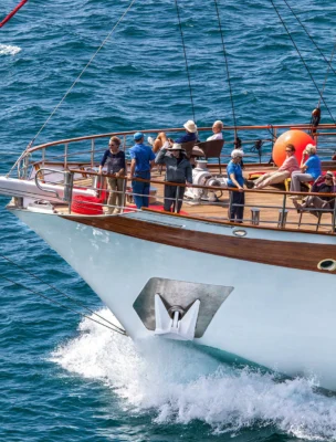 People stand and sit on the deck of a large boat sailing on a sunny day, with water splashing at the front.