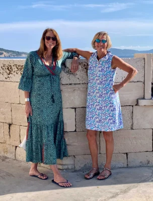 Two women in sunglasses pose next to a stone wall, with a scenic backdrop of mountains and water. One wears a long green dress, the other a blue and white dress with sandals.