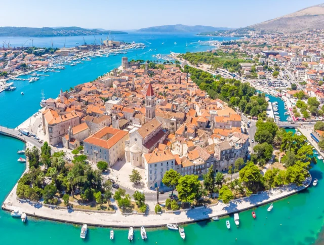 Aerial view of a coastal town with red-roofed buildings, surrounded by turquoise water and boats, with a bridge connecting to the mainland.