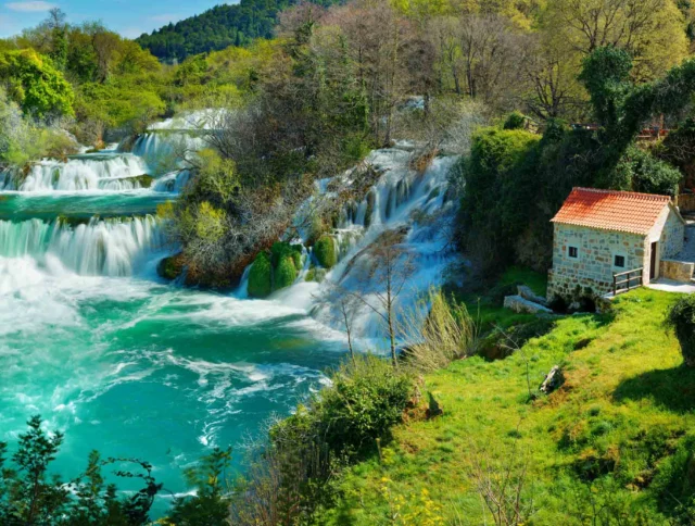 Lush landscape with a waterfall flowing into a turquoise river, surrounded by greenery and two stone houses with red roofs.