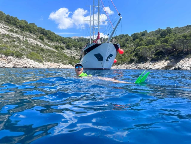 Person swimming near a large sailboat in a clear blue bay, surrounded by rocky hills and trees under a partly cloudy sky.