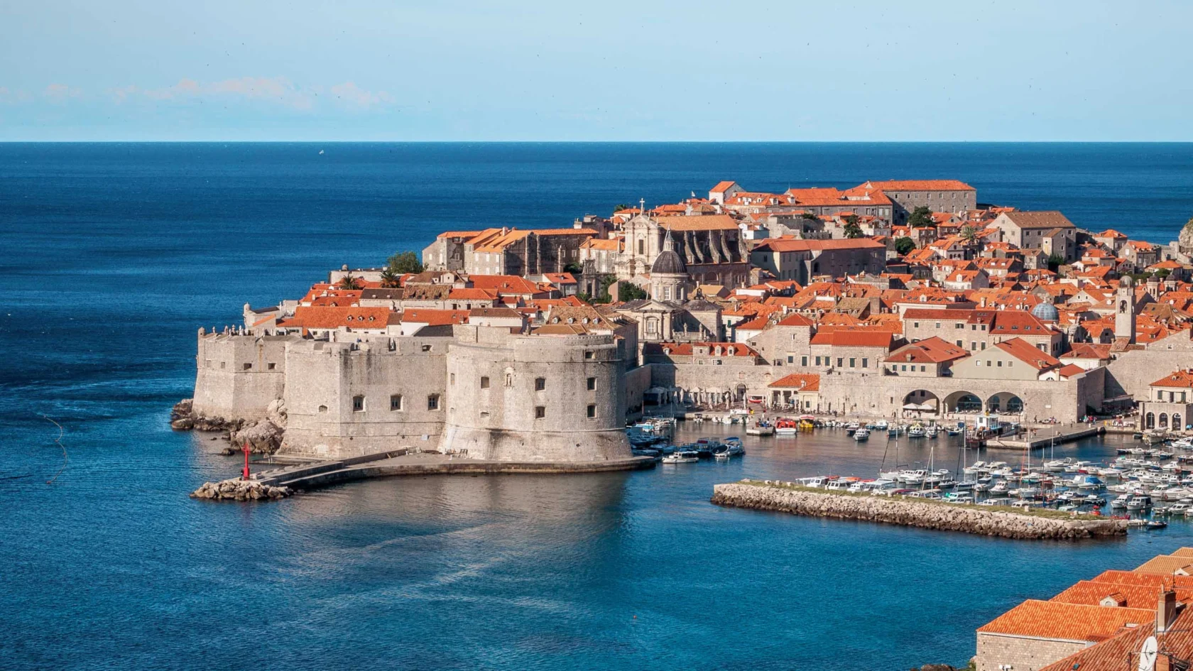 Aerial view of a historic coastal city with stone buildings and orange rooftops, surrounded by blue sea. A small harbor with boats is visible.