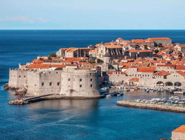 Aerial view of a historic coastal city with stone buildings and orange rooftops, surrounded by blue sea. A small harbor with boats is visible.