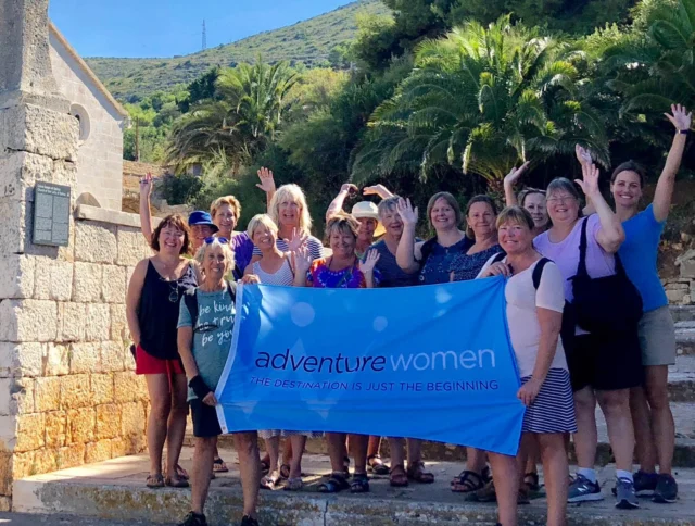 Group of women on stone steps in front of a building and trees, holding a blue "Adventure Women" banner and waving.