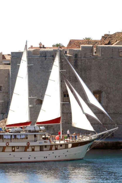 A sailboat with three sails glides near an ancient stone fortress by the sea, under a clear sky.