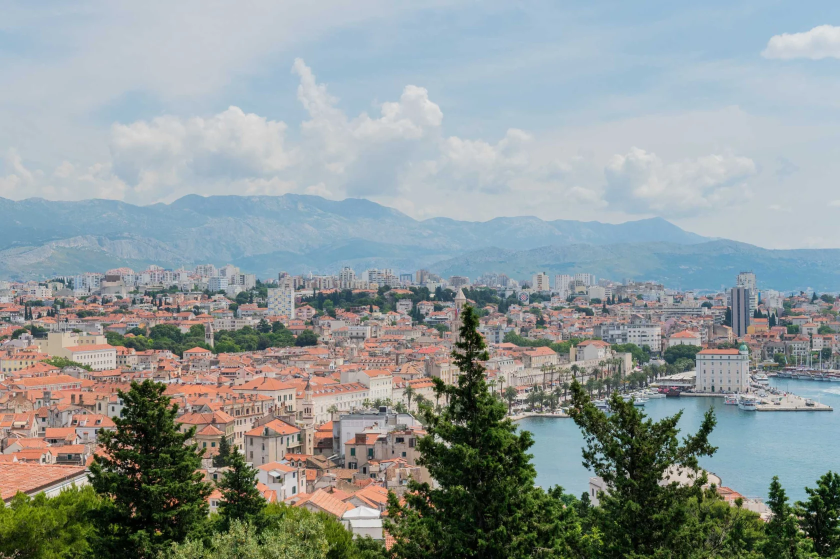 A scenic view of a coastal city with terracotta-roofed buildings, a harbor, and distant mountains under a partly cloudy sky. Green trees are in the foreground.