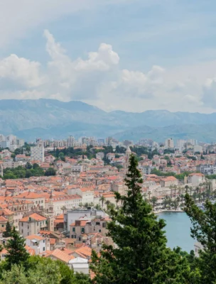 A scenic view of a coastal city with terracotta-roofed buildings, a harbor, and distant mountains under a partly cloudy sky. Green trees are in the foreground.