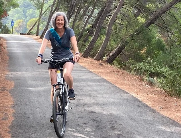 A woman with gray hair rides a bicycle on a paved path through a forested area.