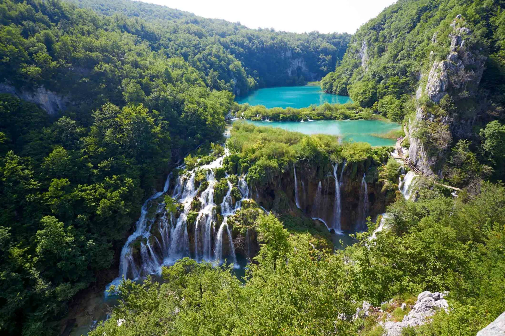 Lush green landscape with cascading waterfalls flowing into turquoise lakes surrounded by forested hills.