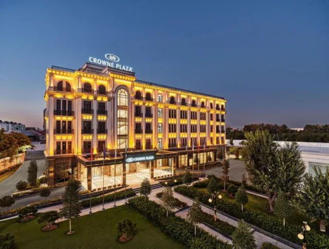 A multi-story Crowne Plaza hotel building is illuminated at dusk, with a landscaped garden, paths, and trees in the foreground.
