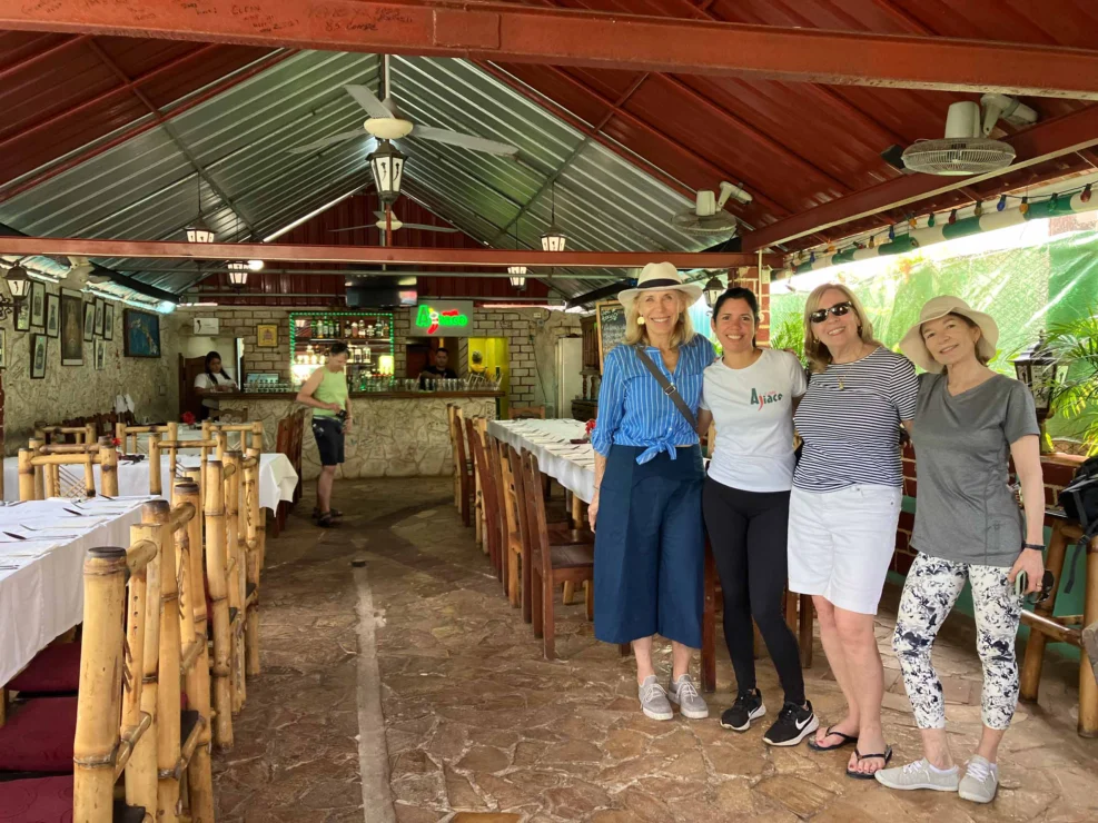 Four women pose together inside a rustic restaurant with a long dining table and a bar in the background.