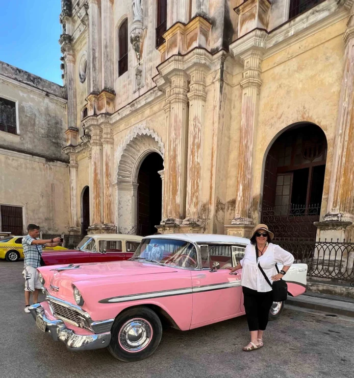 A woman stands beside a pink vintage car in front of an old stone building with ornate columns. A man is visible in the background.