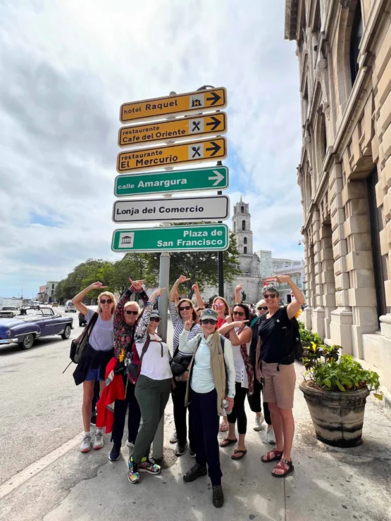 A group of people pose and point at a multi-directional street sign on a city sidewalk, with a historic building and parked cars visible in the background.