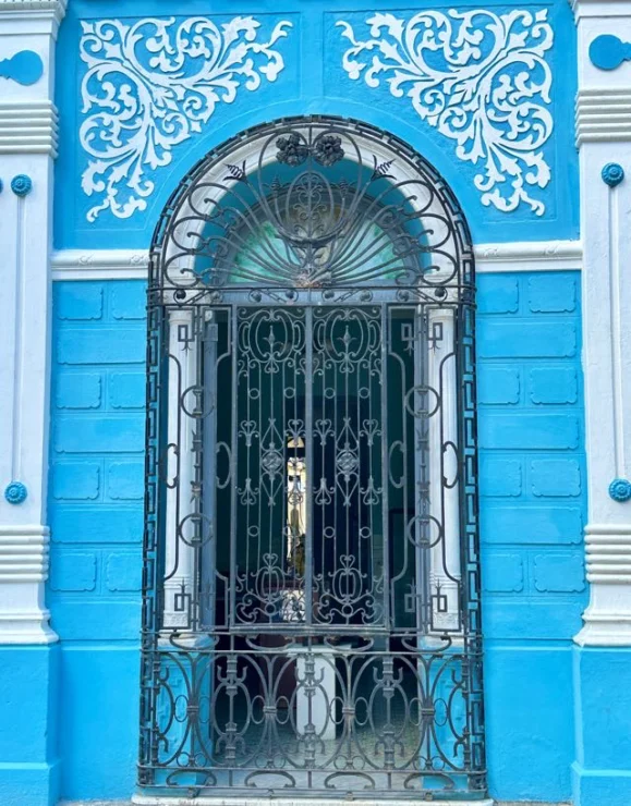 Ornate metal gate on a bright blue building with white decorative patterns around an arched window.