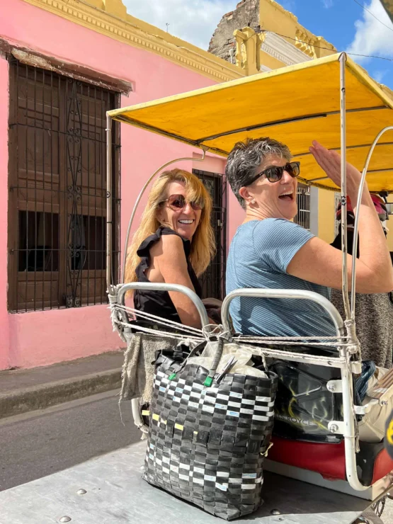 Two people smiling while riding in a vehicle with a yellow canopy. A pink building is in the background.