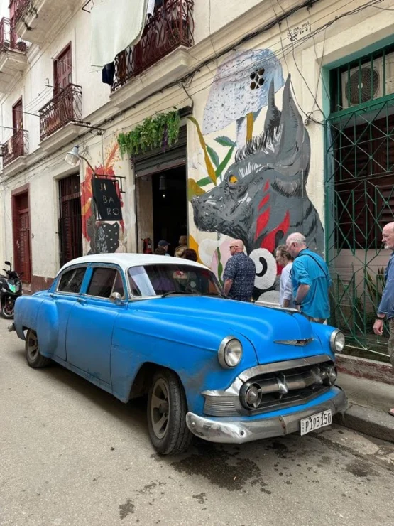 A vintage blue car parked on a street with people nearby, in front of a mural featuring abstract art on a building wall.