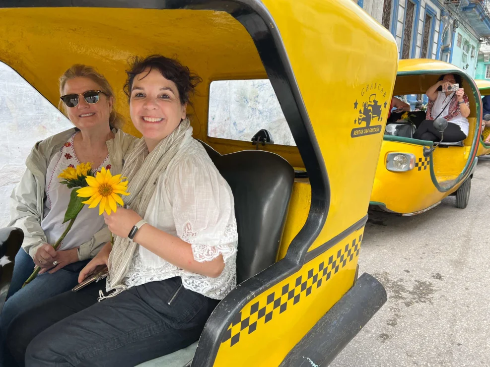 Two women sit in a yellow three-wheeled taxi, one holding a sunflower. Another person is seated in a similar taxi behind them.