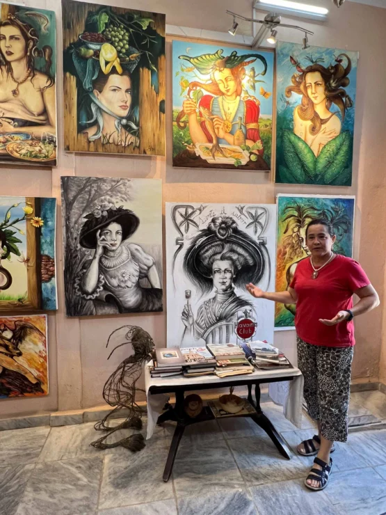 Person in a red shirt standing in an art gallery, gesturing toward a wall display of colorful paintings and one black-and-white drawing. A table with books is in front.