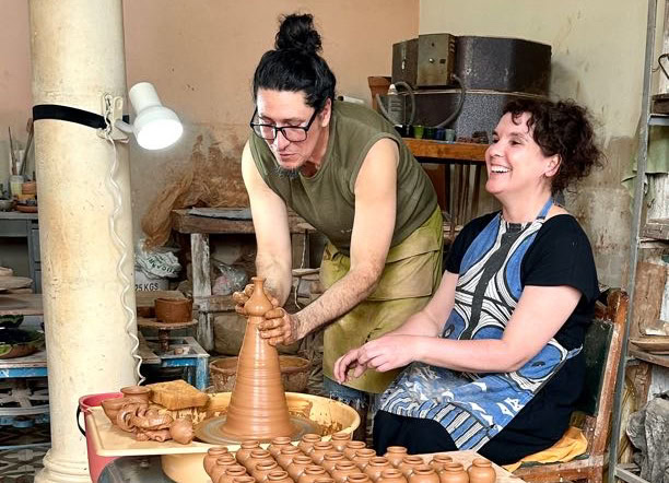 A person demonstrates pottery making on a wheel while another person sits nearby, smiling. The room is filled with pottery tools and finished pieces.