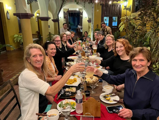 A group of people seated at a long dining table, raising glasses in a toast, surrounded by plates of food and drinks in a warmly lit room.