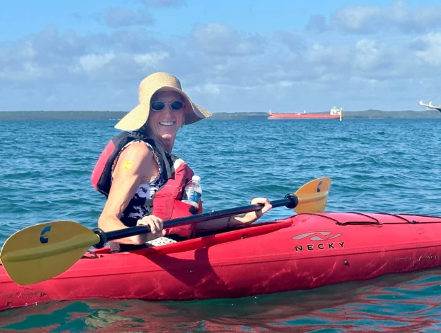 A person in a sun hat and life jacket is kayaking on open water with ships in the background on a sunny day.