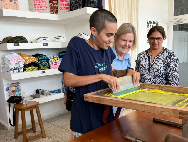 Person screen printing with guidance in a shop, featuring stacked printed items and two onlookers in the background.