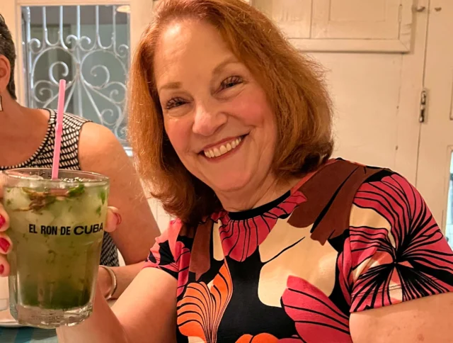 A smiling person holds a drink with a straw in a glass labeled "El Ron de Cuba." They are wearing a floral-patterned shirt.