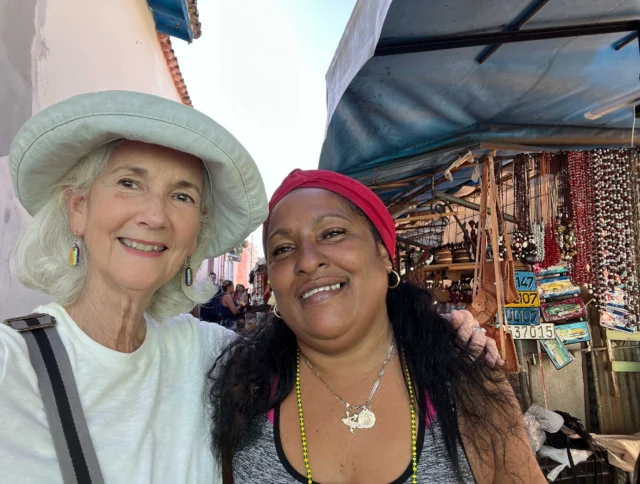 Two women smiling together in an outdoor market, with stalls displaying various items in the background. One wears a white hat, and the other a red headscarf.