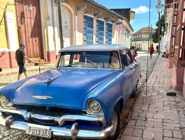 A vintage blue car is parked on a cobblestone street lined with colorful colonial buildings under a clear blue sky.